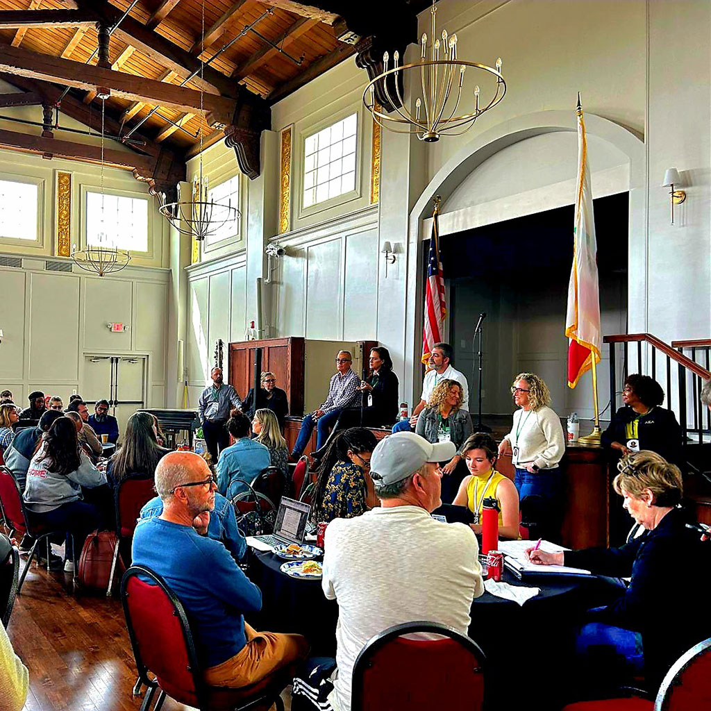 People sit at round tables while a group speaks on stage in a large, sunlit hall with high ceilings and chandeliers.