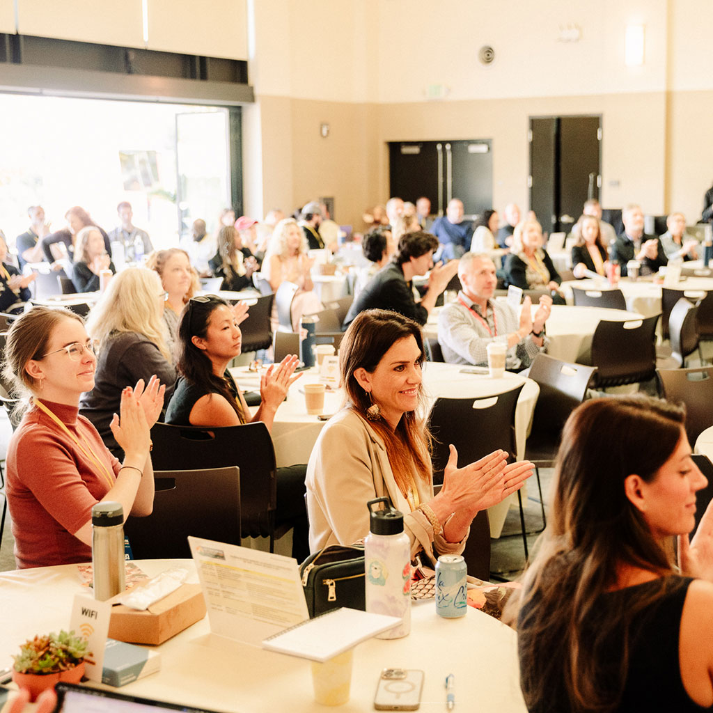People seated at round tables in a bright room, attentively listening and applauding a speaker discussing programs that support community to community initiatives.