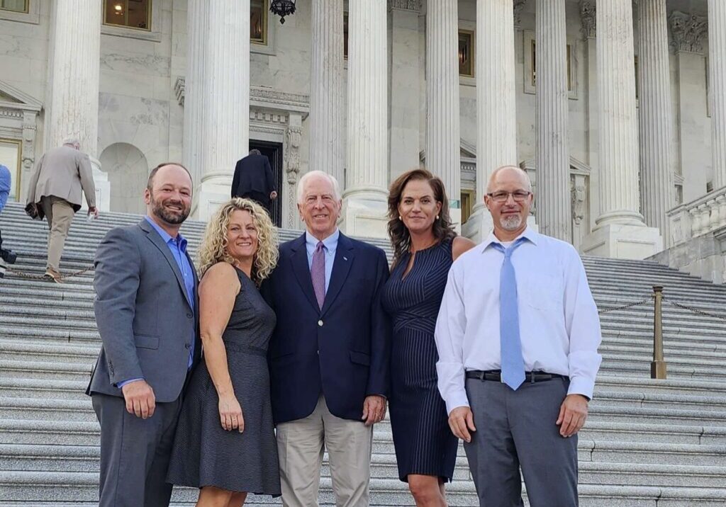 Five people pose and smile on the steps of a government building with white columns in the background, celebrating community recovery with After the Fire USA.