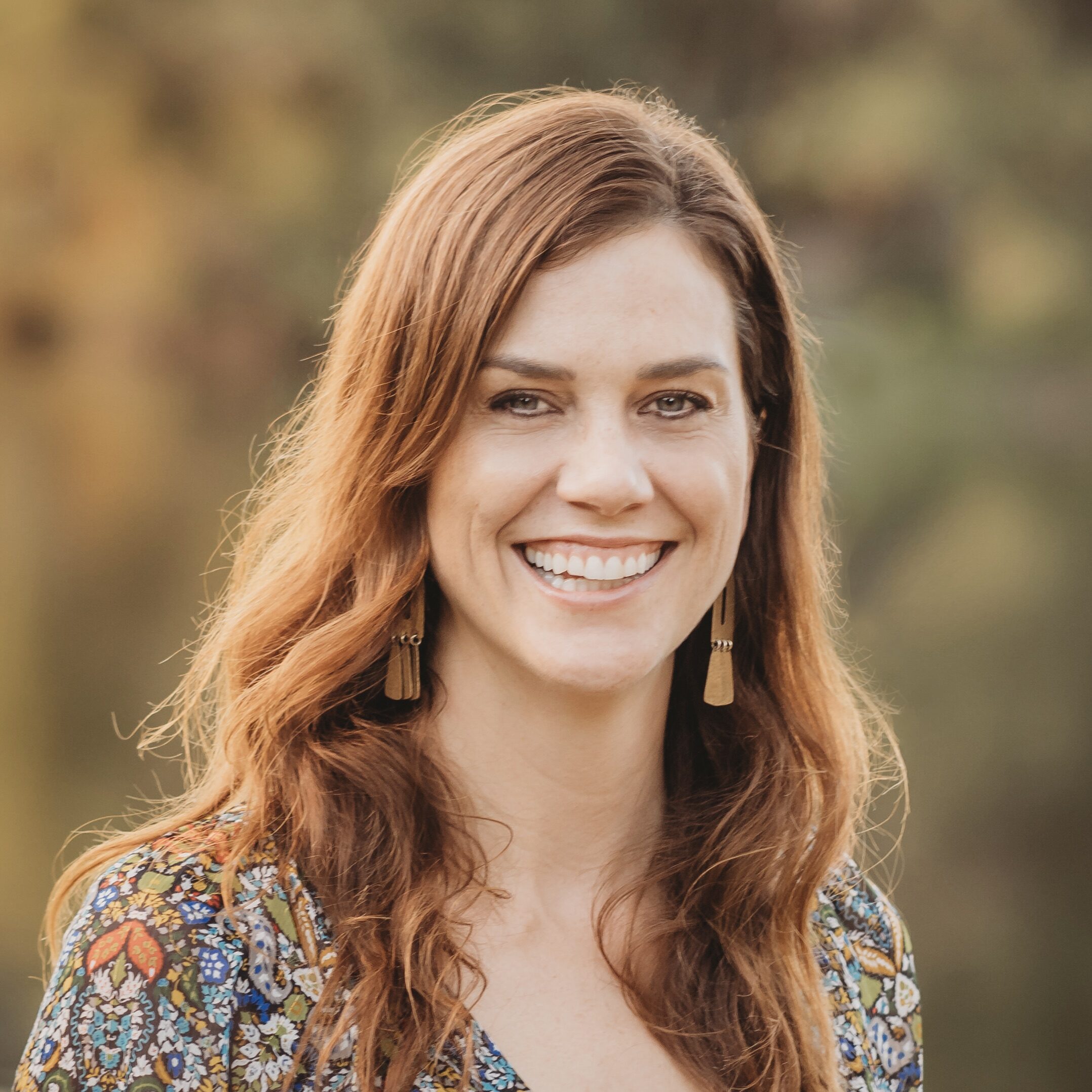 Woman with long reddish-brown hair smiling outdoors, wearing patterned top and gold earrings.
