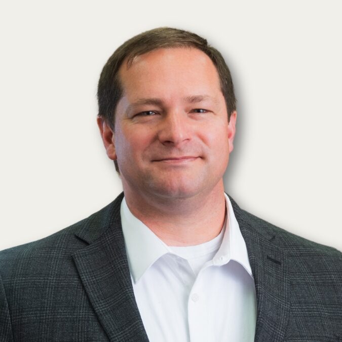 Man in a gray blazer and white shirt smiling at the camera against a plain background.