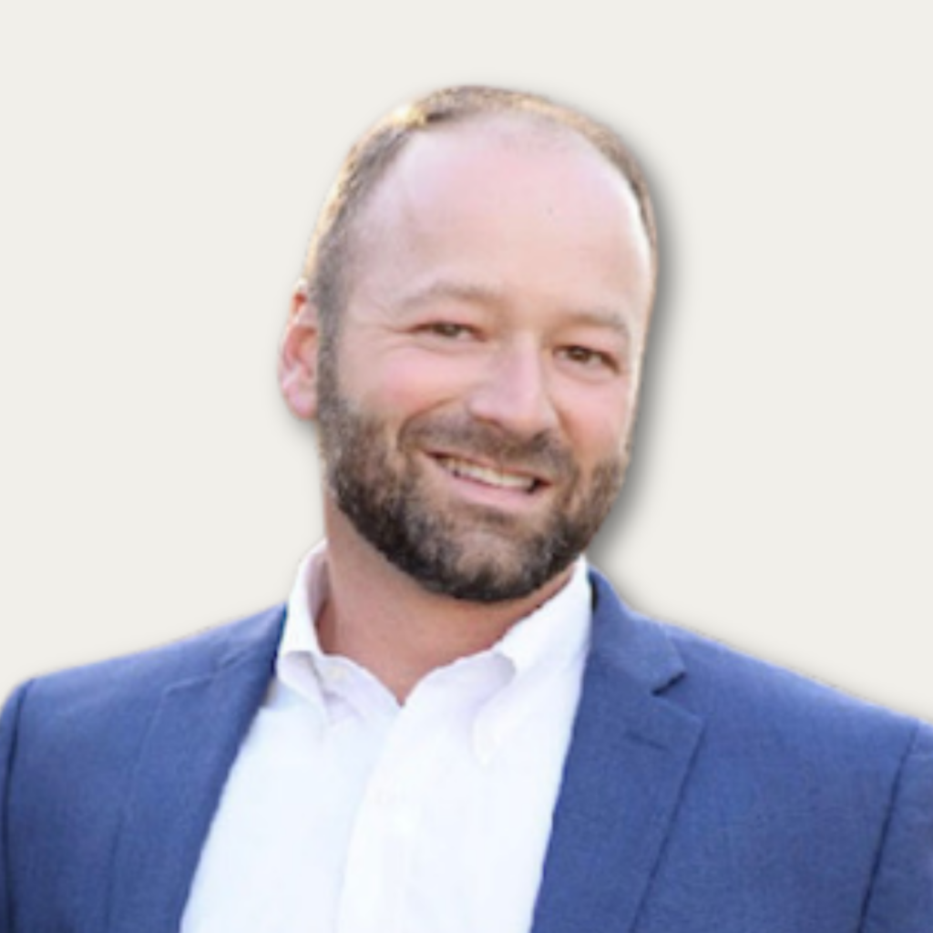 Smiling man with a beard wearing a blue suit jacket and white shirt against a light background.