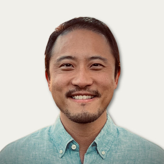 Smiling man with short dark hair and a trimmed beard, wearing a light blue button-up shirt, on a white background.