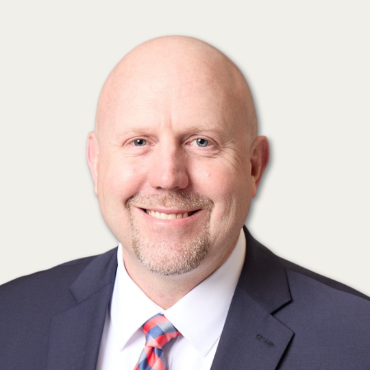 Bald man in a suit and striped tie smiling in front of a plain light background.