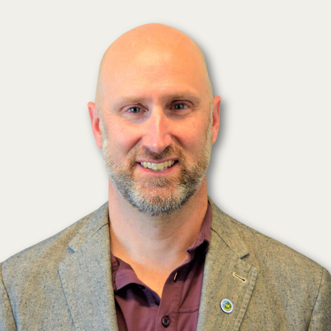 Bald man with a gray beard wearing a gray blazer and maroon shirt, smiling at the camera.