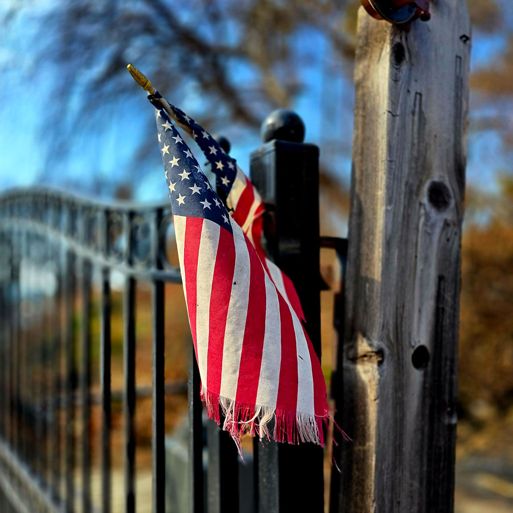 A slightly worn American flag hangs on a metal fence near a wooden post, with trees in the background.