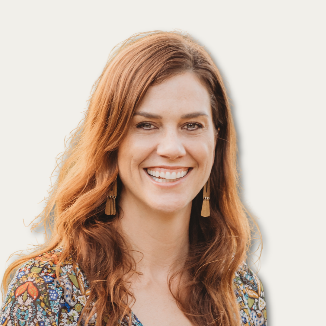 Woman with long red hair smiling, wearing a patterned top and tasseled earrings, against a white background.