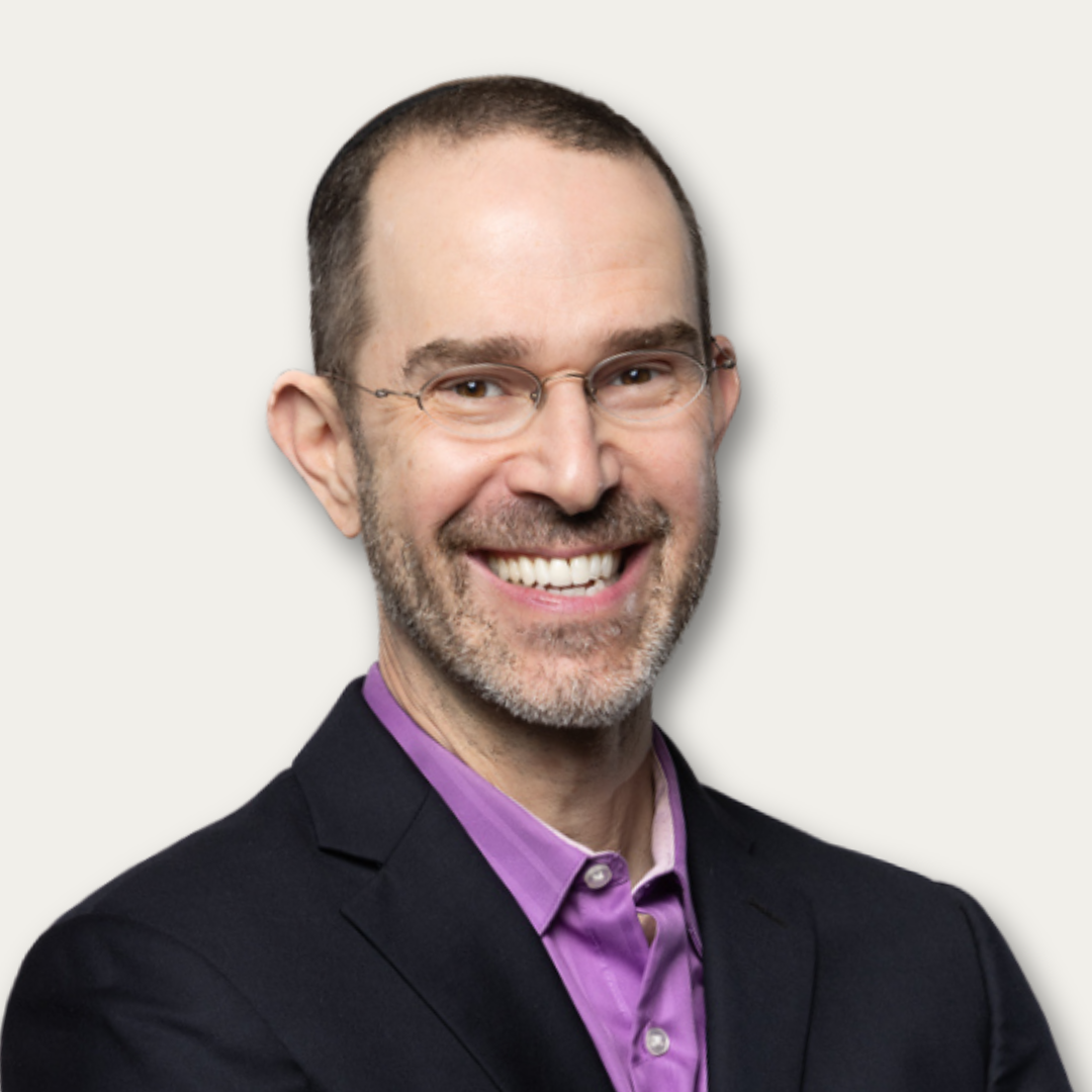 Smiling man with glasses, short hair, and beard wearing a purple shirt and dark blazer against a light background.