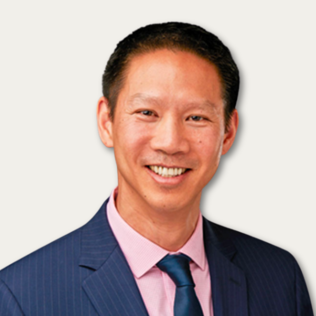 Smiling man in a blue suit, pink shirt, and blue tie, facing forward against a plain light background.