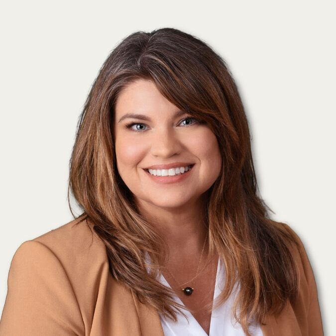 Smiling woman with long brown hair in a tan blazer and white shirt, posing against a light background.