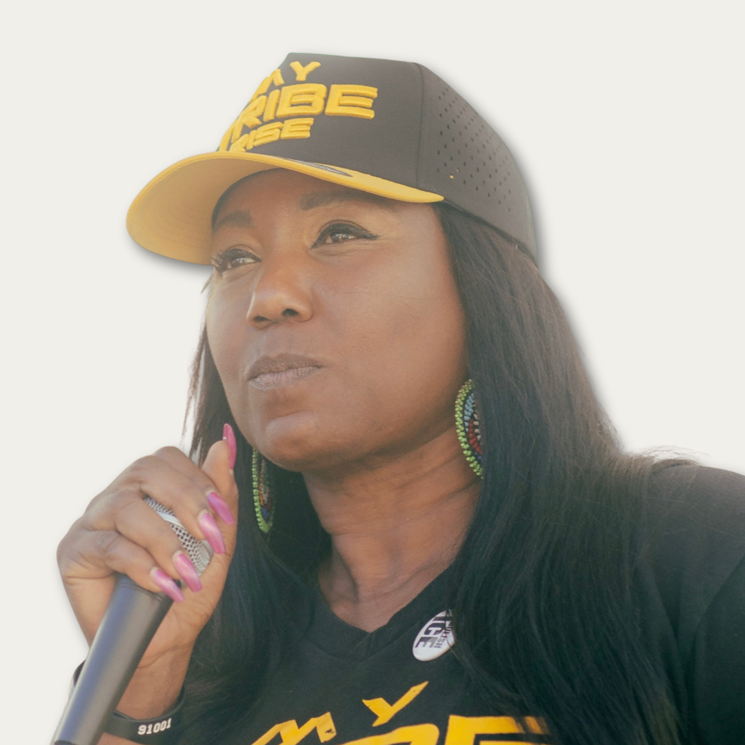 Woman in a black cap holding a microphone, wearing large earrings and a black shirt with yellow text.