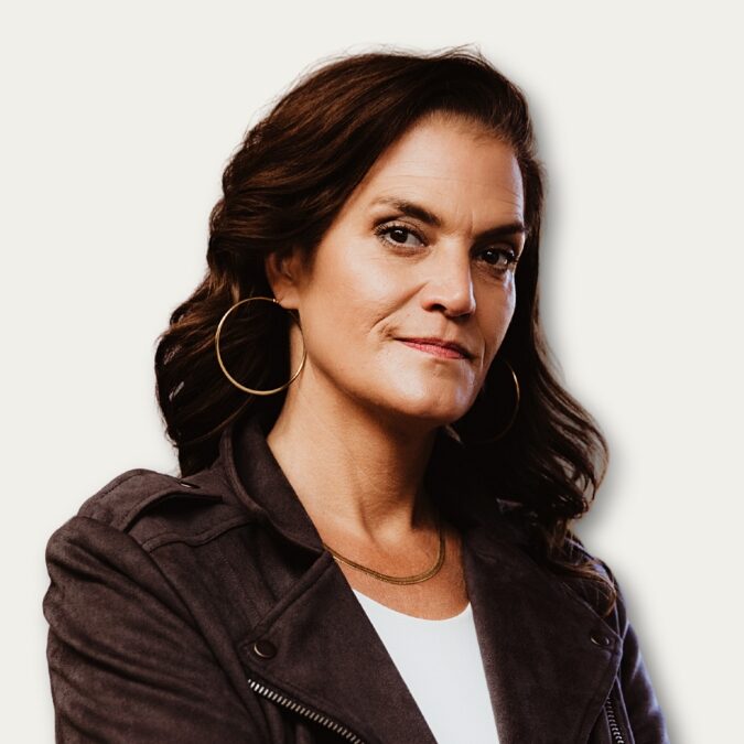 Woman with wavy brown hair in a dark jacket, large hoop earrings, and a white top, looking confident.