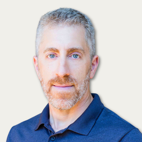 Man with short gray hair and beard wearing a dark polo shirt, facing camera against light background.
