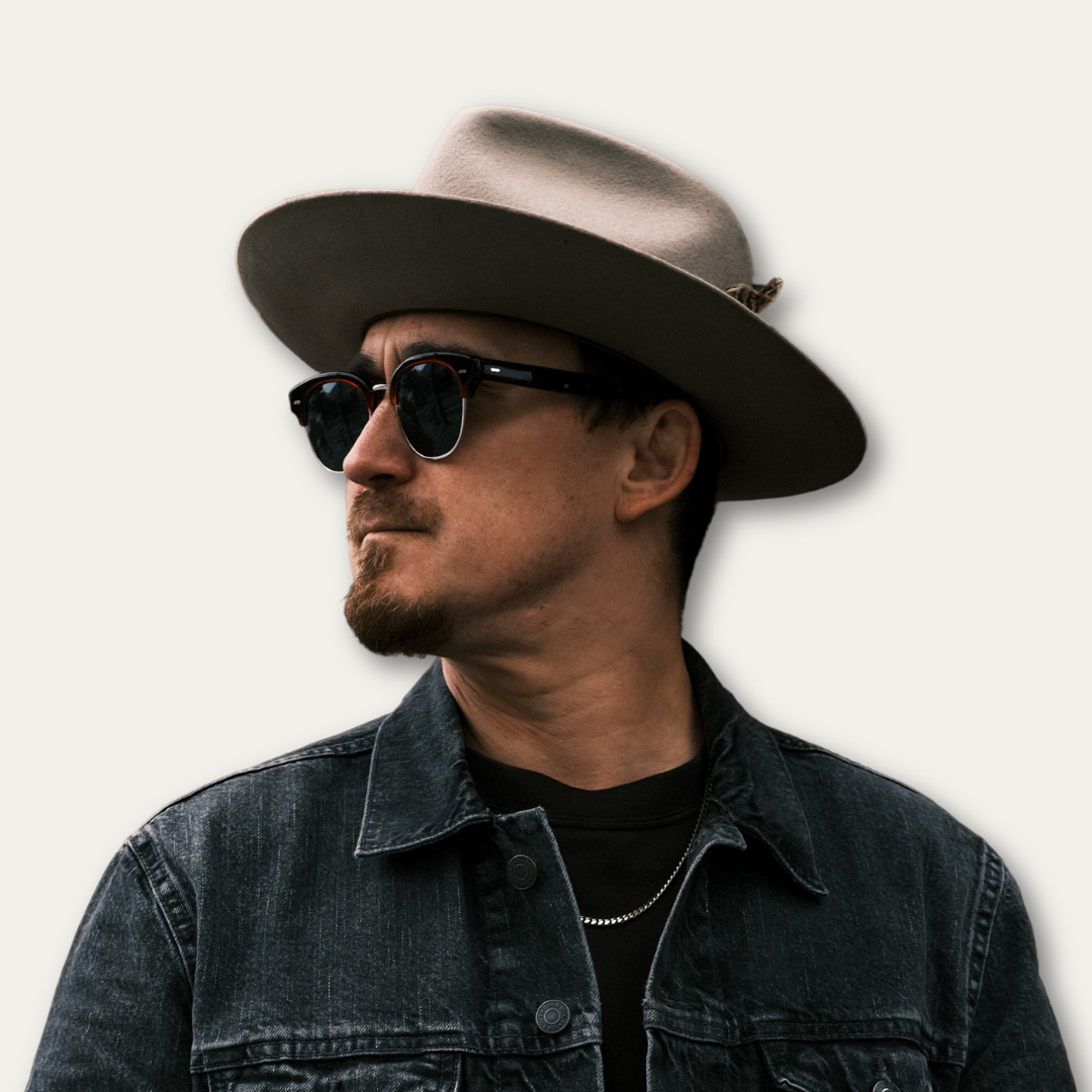 Man in sunglasses and a wide-brimmed hat, wearing a denim jacket, looking to the side.