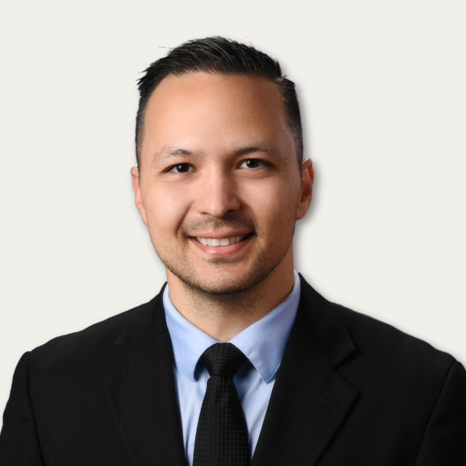 Smiling man in a suit and tie with short dark hair against a plain light background.