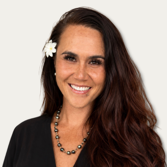 Smiling woman with long brown hair, a white flower in her hair, and a black necklace, wearing a black top.