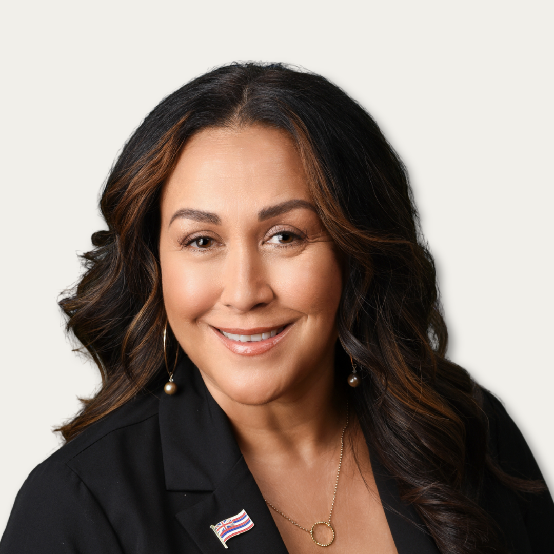 Woman with long dark hair, wearing a black blazer, necklace, and flag pin, smiling at the camera.