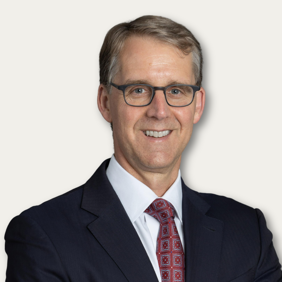 Smiling man in a suit and patterned tie with glasses, standing against a plain, light-colored background.