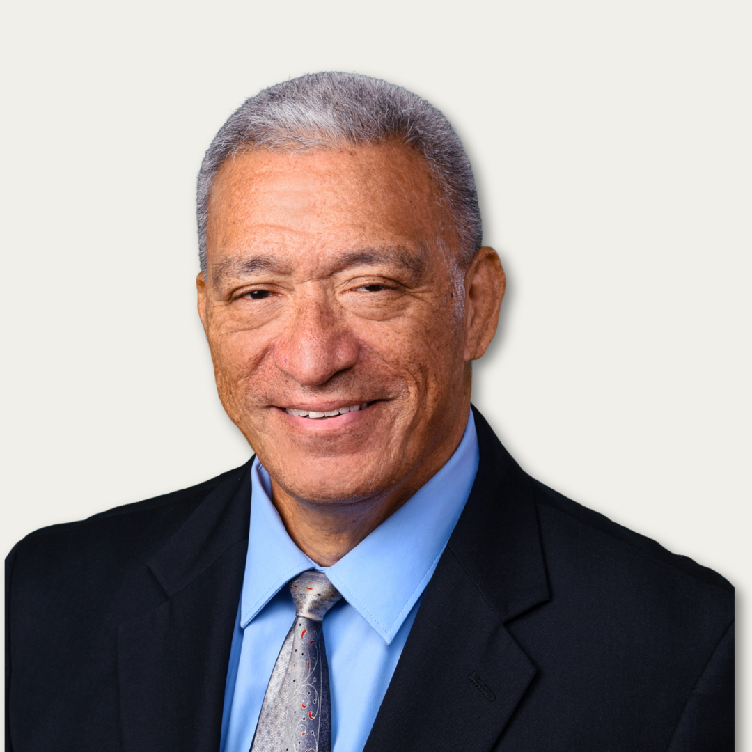Smiling older man with short gray hair in a suit, blue shirt, and tie, on a plain light background.