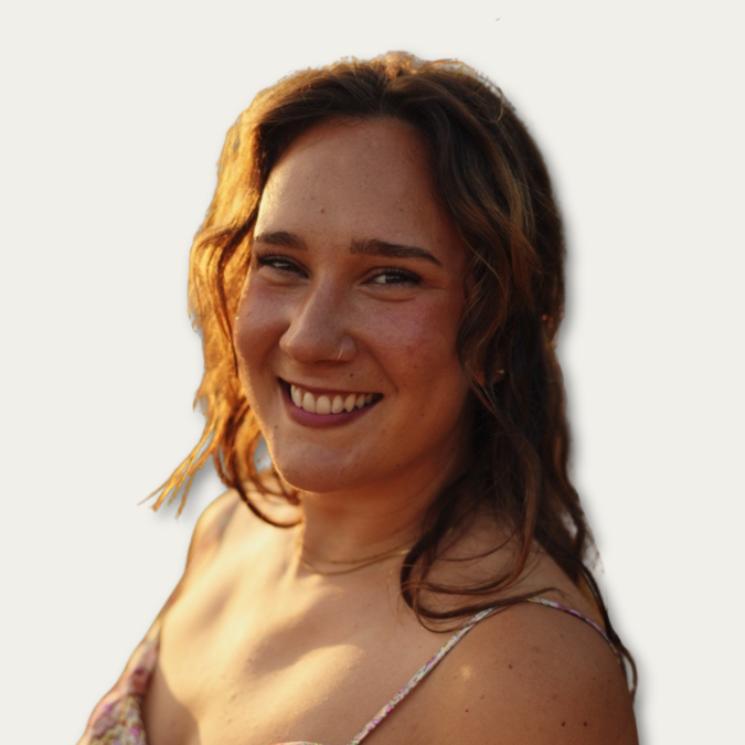 Woman with wavy brown hair smiling at the camera, wearing a floral dress, on a plain light background.