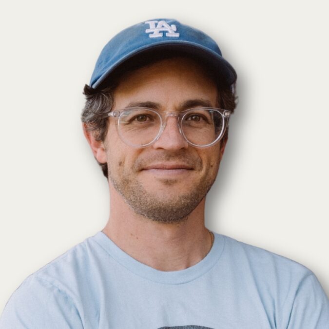 Man with glasses and blue LA cap smiling, wearing a light blue T-shirt, on a plain background.