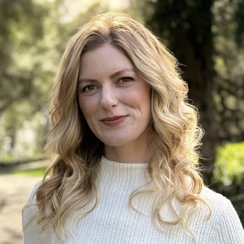 A woman with wavy blonde hair in a white sweater smiles outdoors, embodying the optimism often seen among national leaders supporting fire affected communities, with trees in the background.