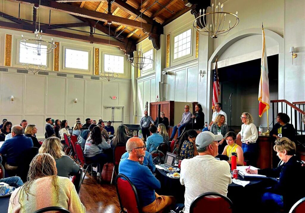 People sit and listen to a panel of speakers in a large, bright room with high ceilings and tall windows, discussing community recovery with After the Fire USA.