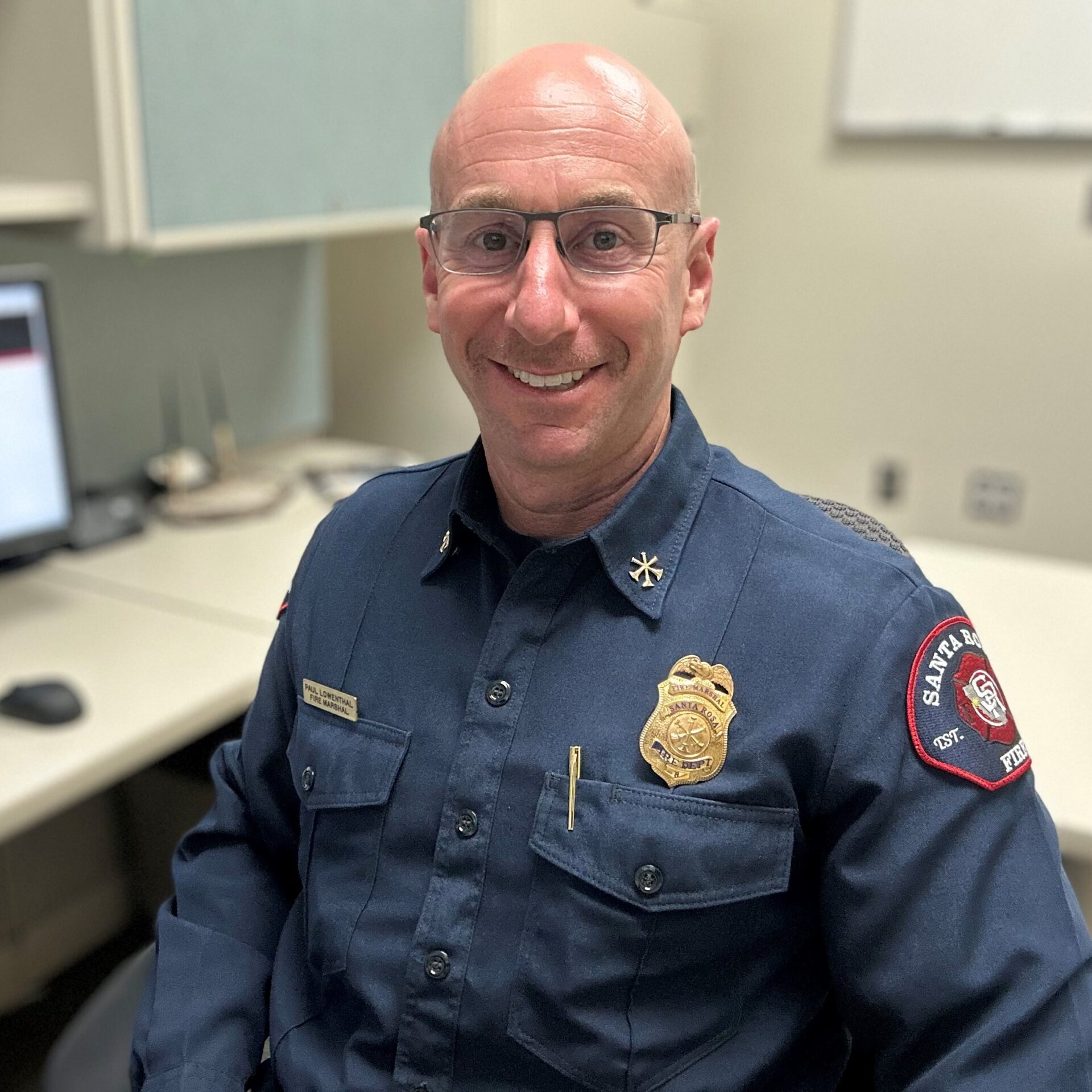Bald man in a fire department uniform smiling while seated at an office desk with a computer.