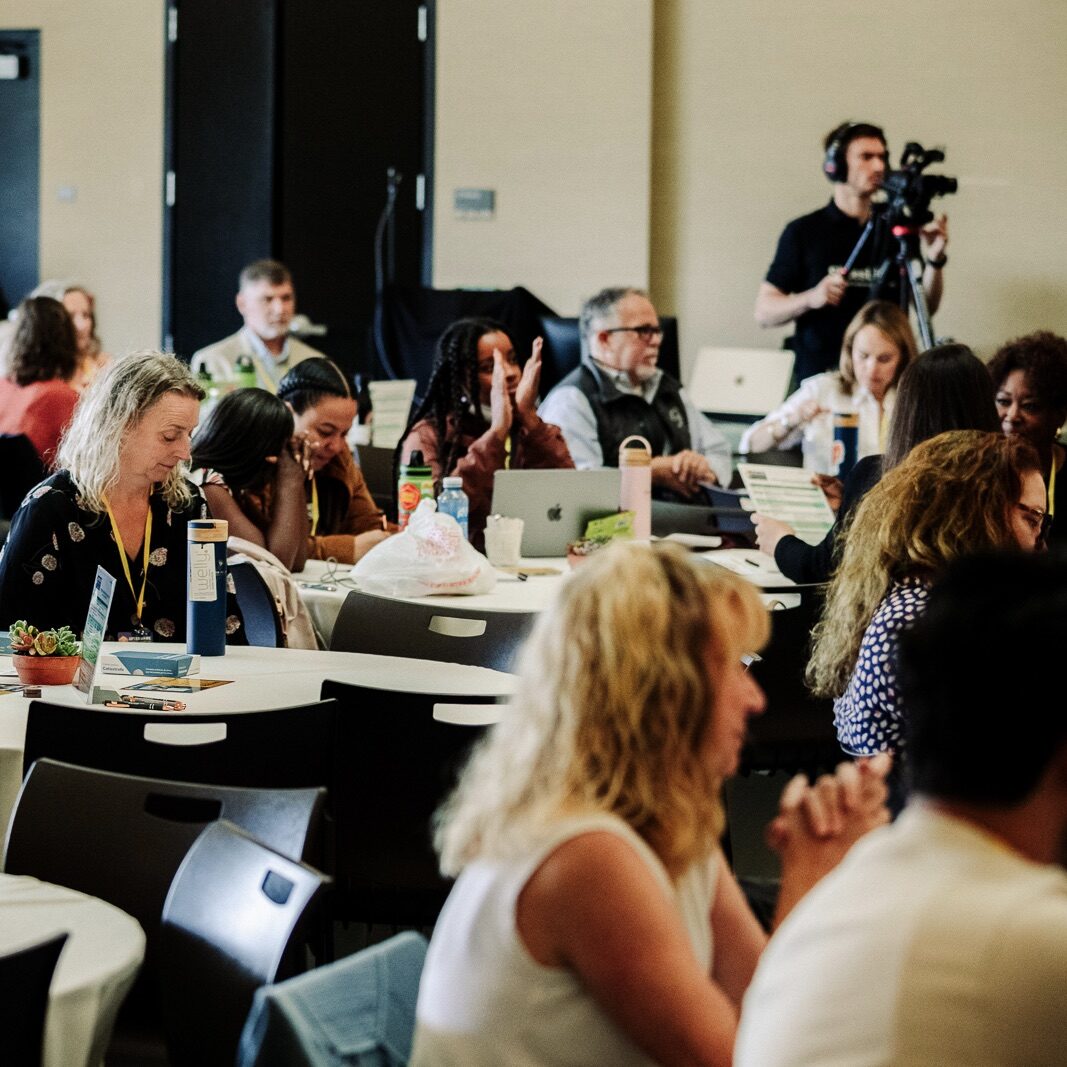 People seated at round tables in a conference room, some taking notes, with a cameraman recording in back.