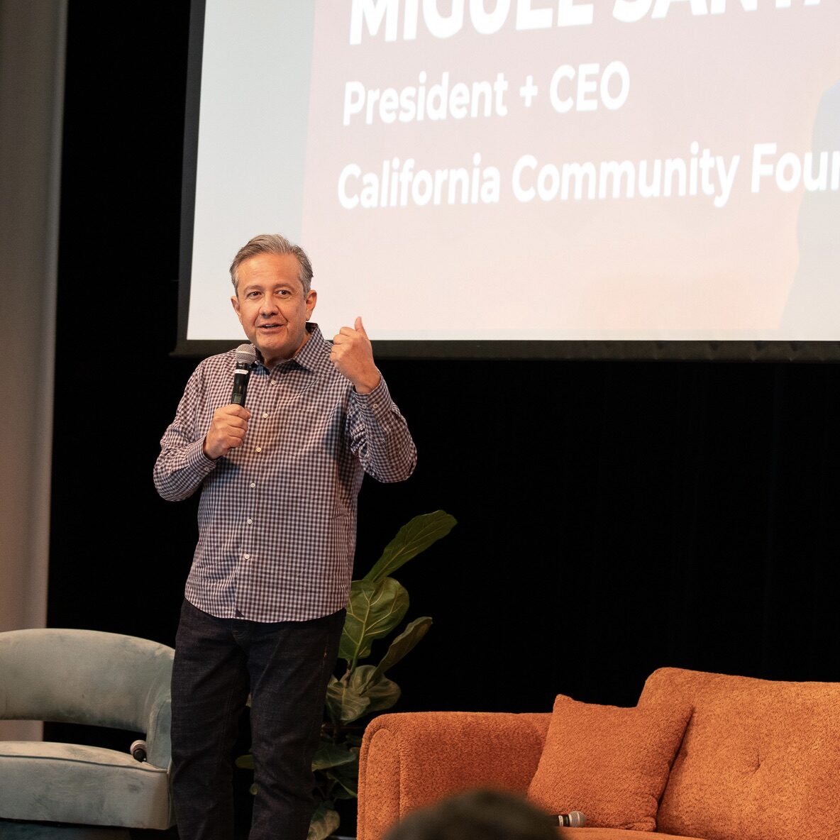 Man speaking on stage with a microphone, standing near a screen displaying text and next to a brown couch.