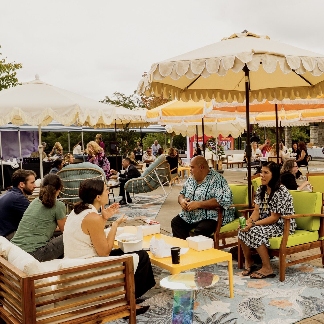 People sit and talk on outdoor lounge furniture under umbrellas at a lively social event.