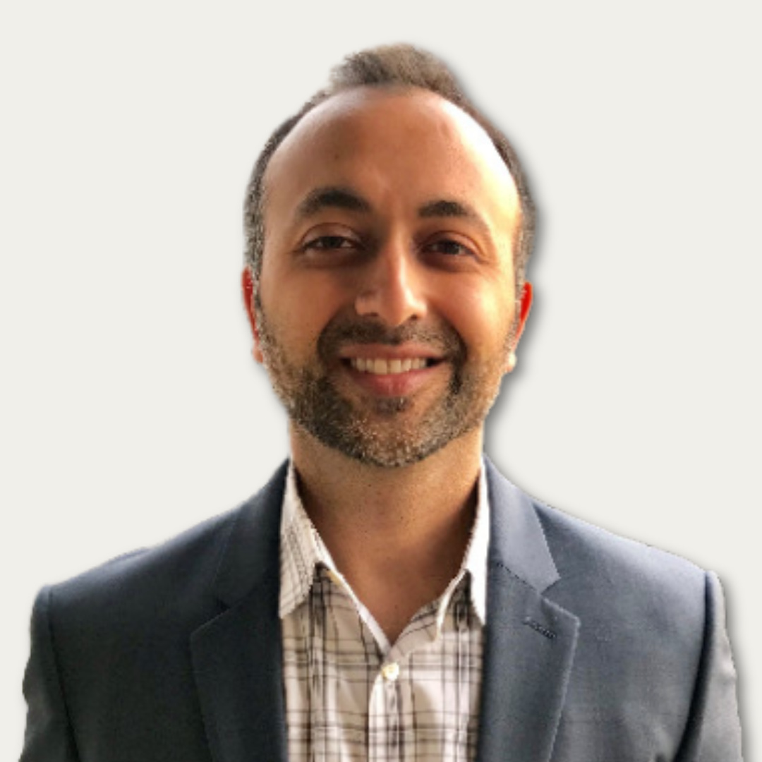 Smiling man with short hair and beard wearing a gray suit jacket and checked shirt on a plain background.
