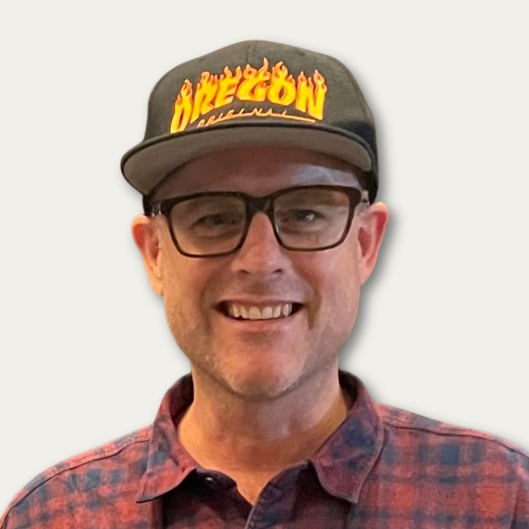 Man in glasses and a plaid shirt wearing a black Oregon hat, smiling against a plain background.