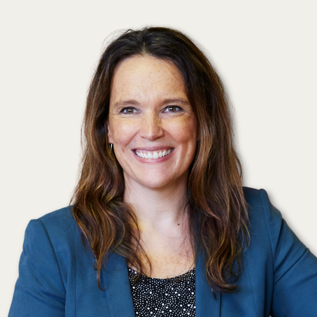 Woman with long brown hair in a teal blazer smiling against a plain light background.