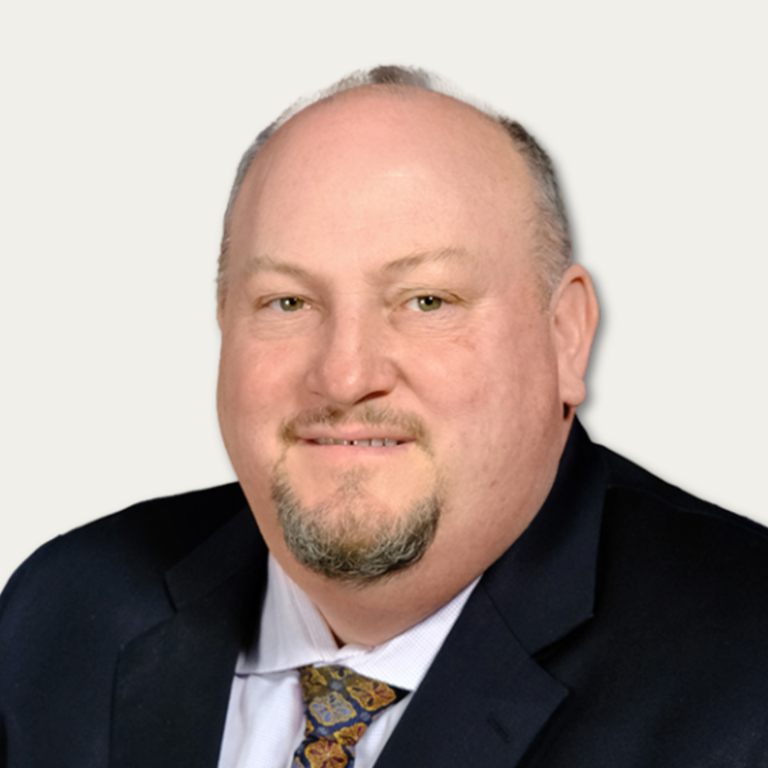 Middle-aged man in a suit and patterned tie smiling at the camera against a plain background.