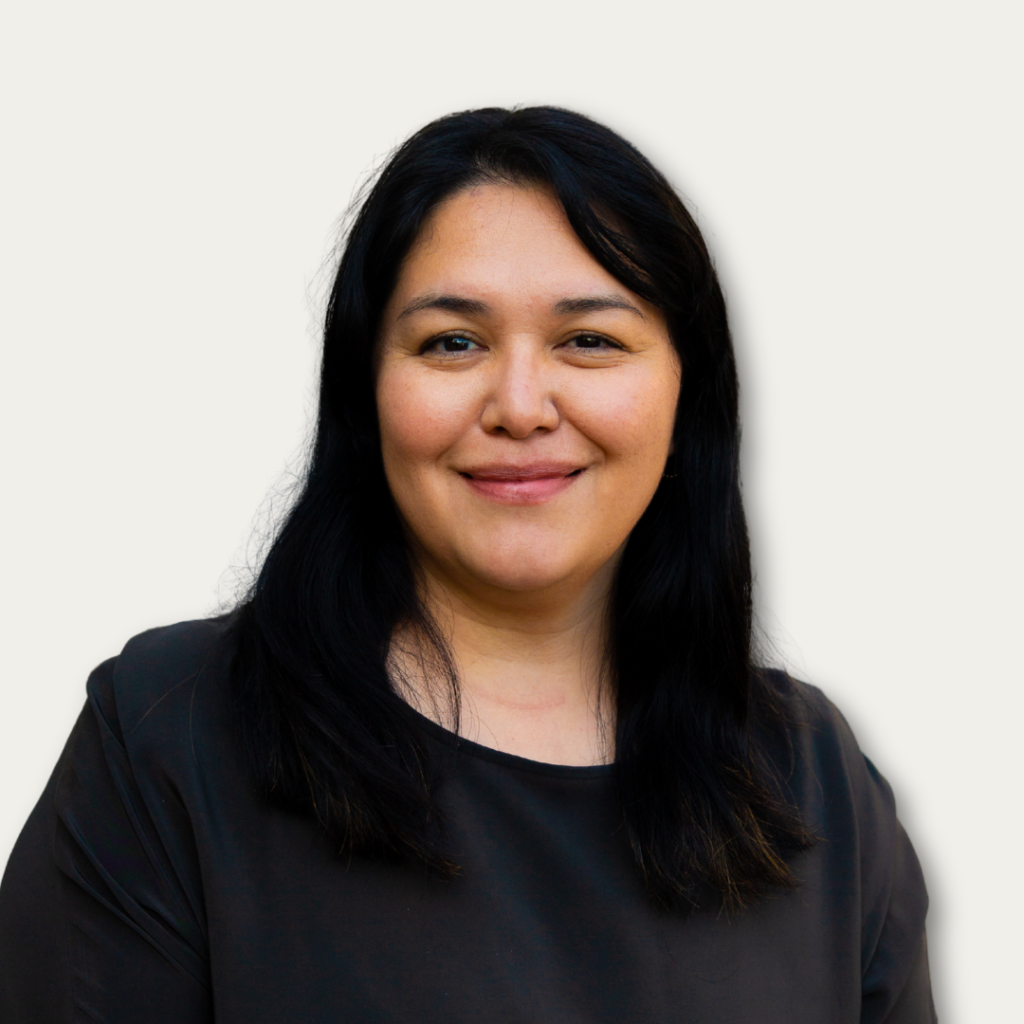 Woman with long dark hair, smiling, wearing a dark top, standing against a plain light background.