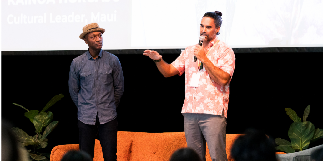 Two men stand on stage speaking; one holds a mic, the other listens. Audience and plants visible.