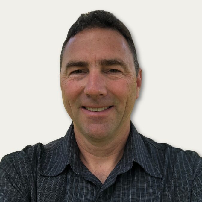 Smiling man with short dark hair wearing a dark, collared shirt, standing against a plain light background.