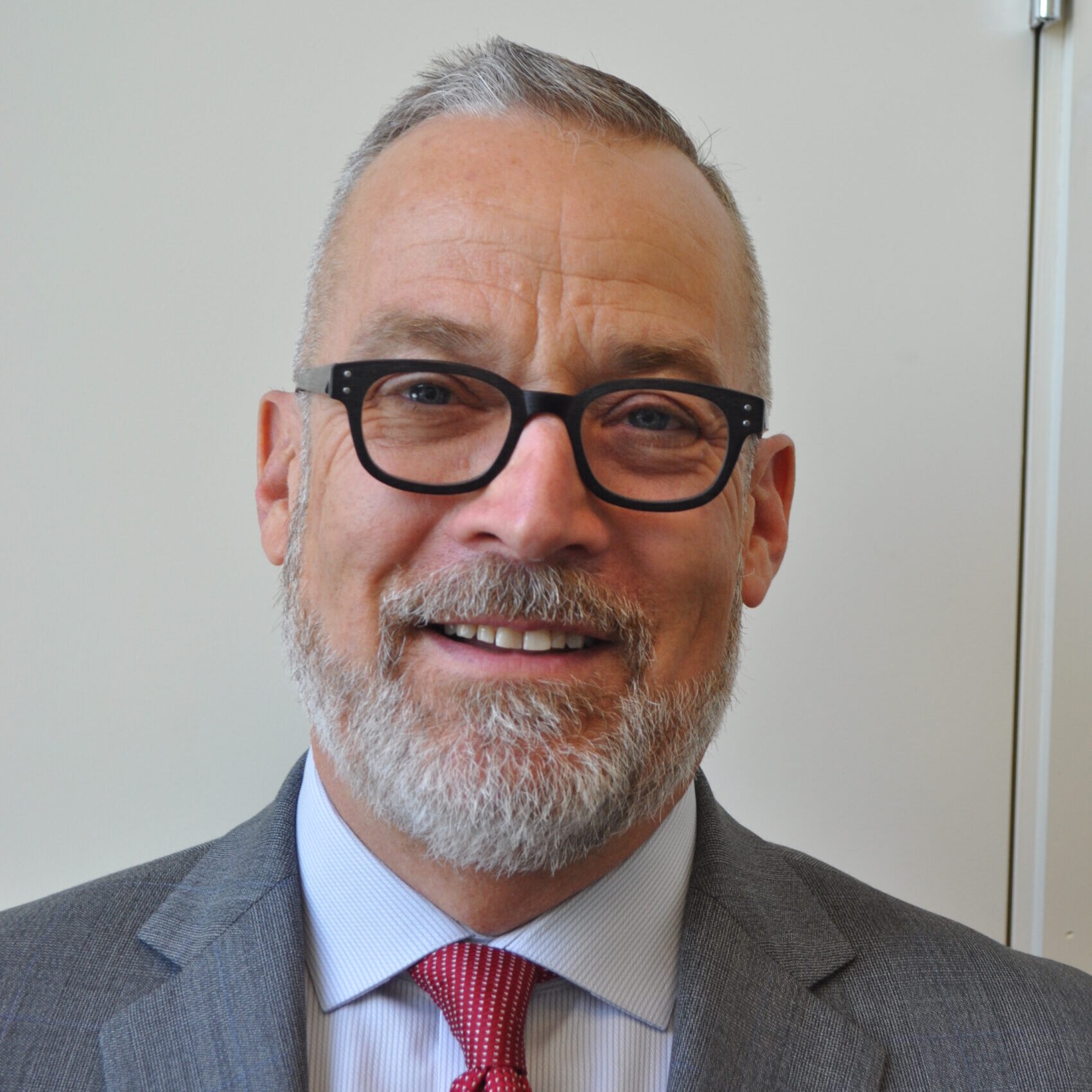 Smiling man with glasses, gray beard, and suit with red tie standing in front of a plain light background.