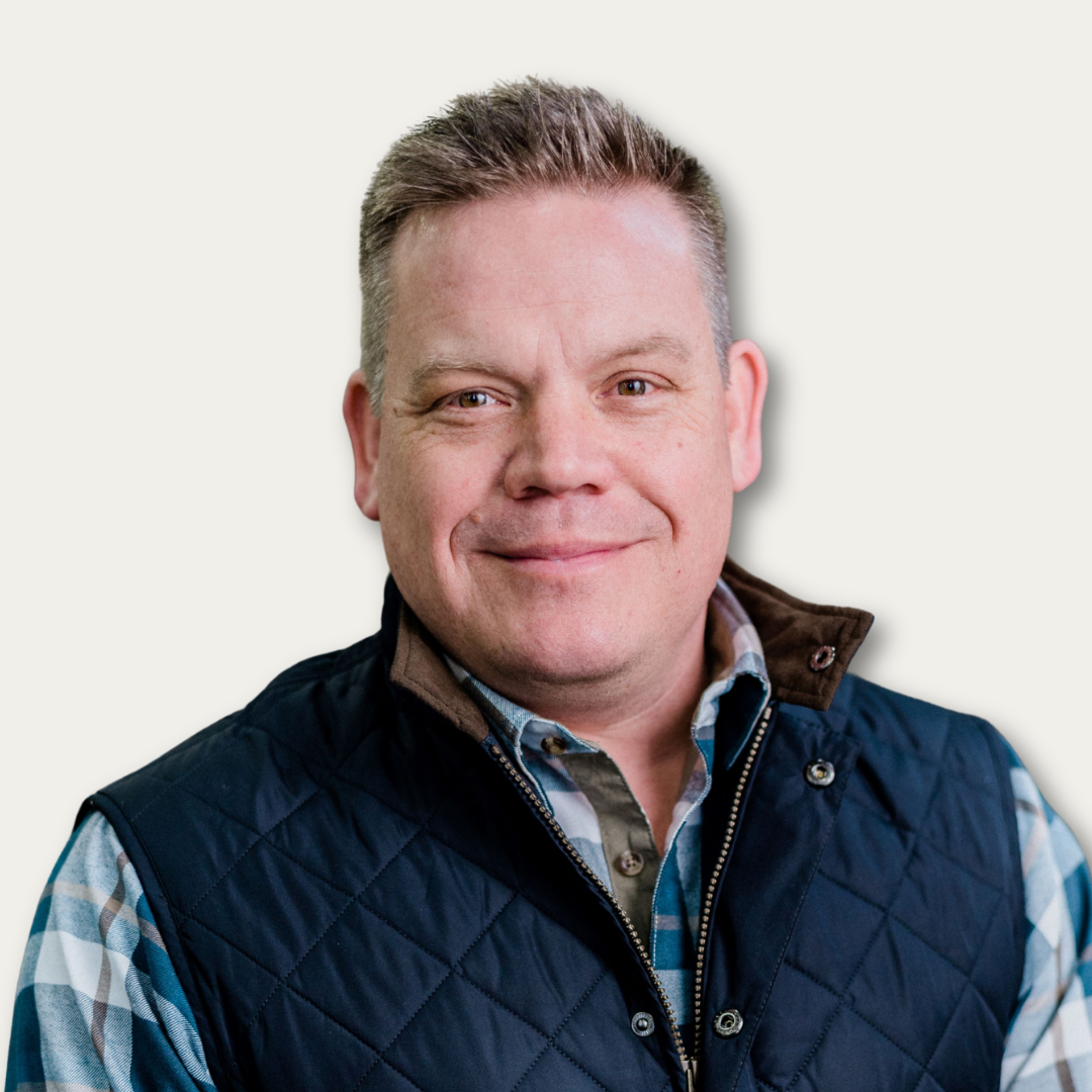 Smiling man in a plaid shirt and quilted vest, standing against a plain, light background.