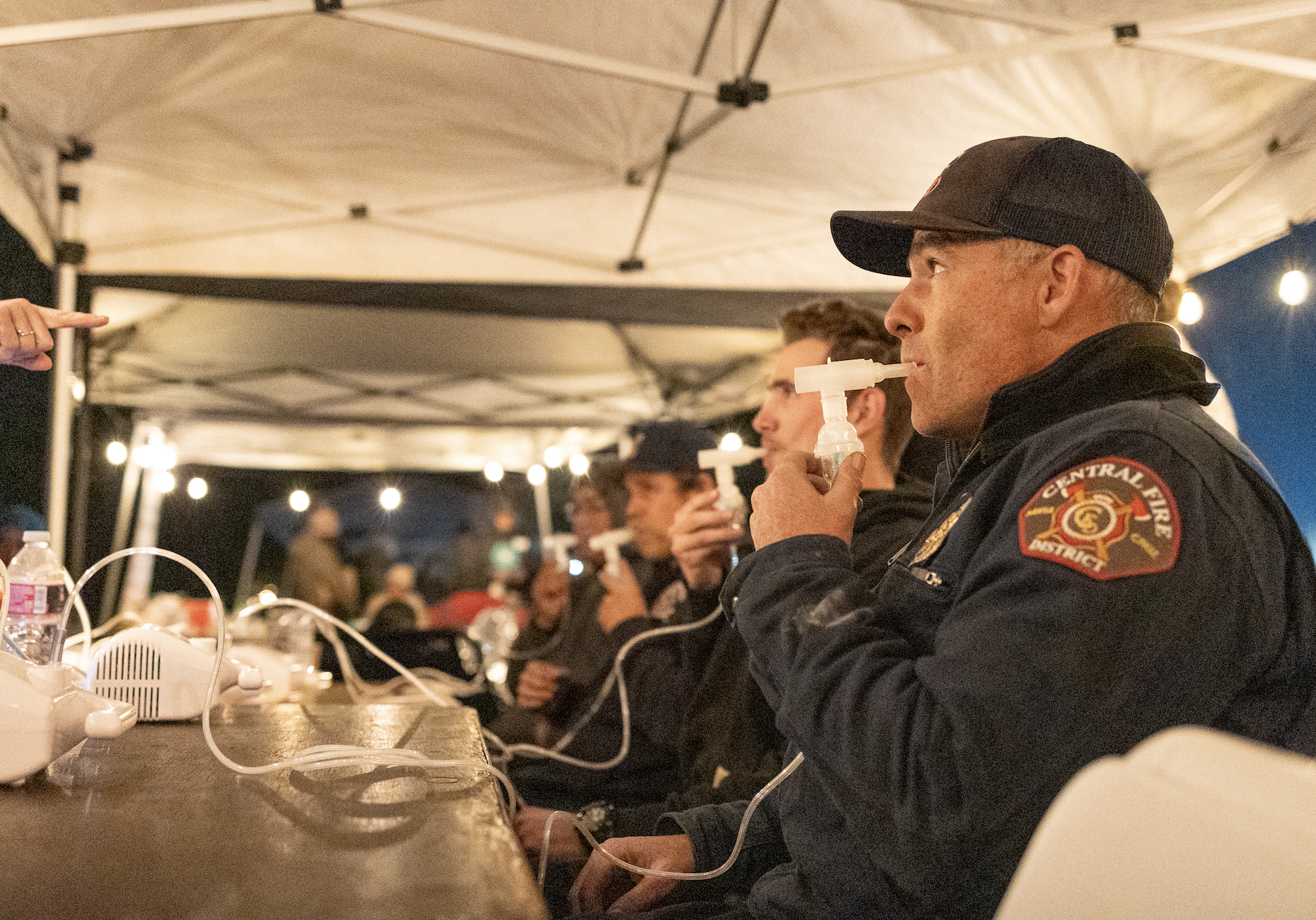 Firefighter in uniform uses a breathing device at a table under a tent with other people.